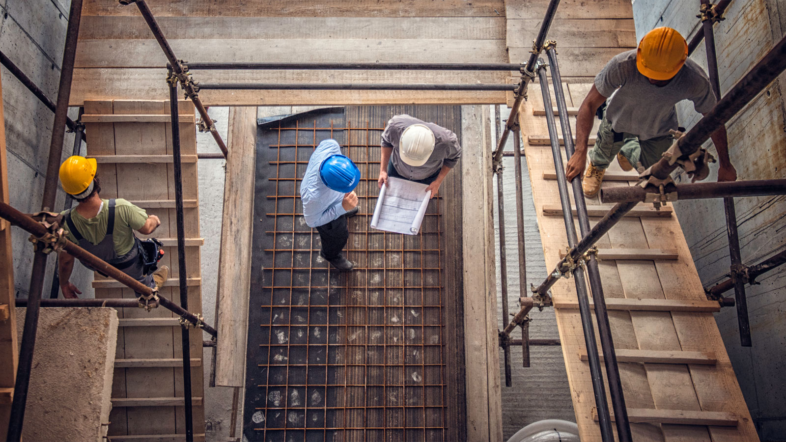 Top view of an architect and an engineer wearing safety helmets looking at blueprints in the middle of a construction site, surrounded by construction workers and building materials.